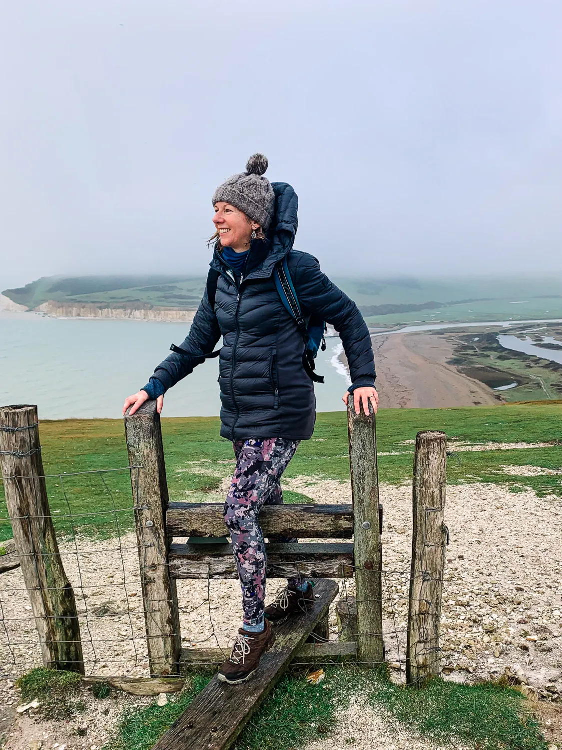 Woman wearing a puffer jacket and knit hat crosses a wooden stile on a coastal hillside, smiling at the sea cliffs in the distance, overcast day.