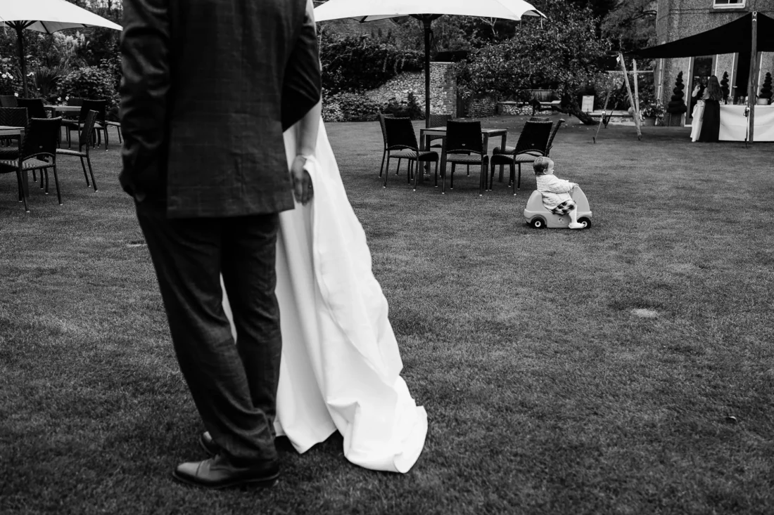 Humourous street-style image in black & white of toddler riding a toy car at Pangdean Barn wedding as bride and groom walk in the opposite direction