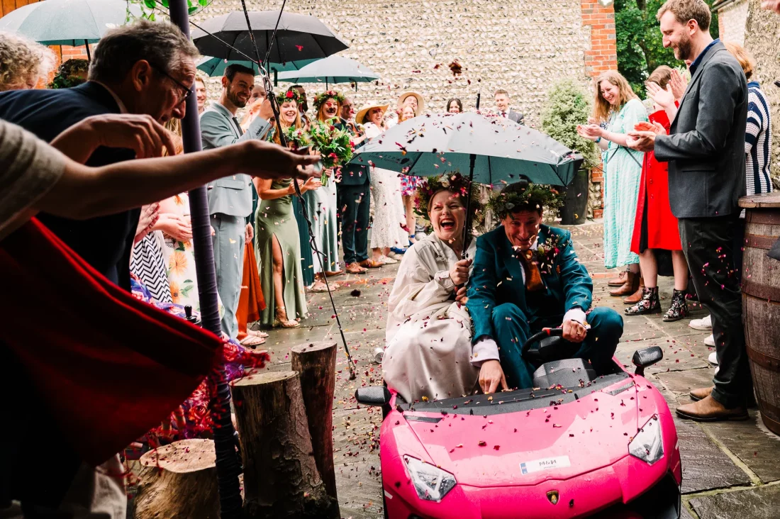 Bride and groom drive through confetti in a pink toy car at Cissbury Barns wedding