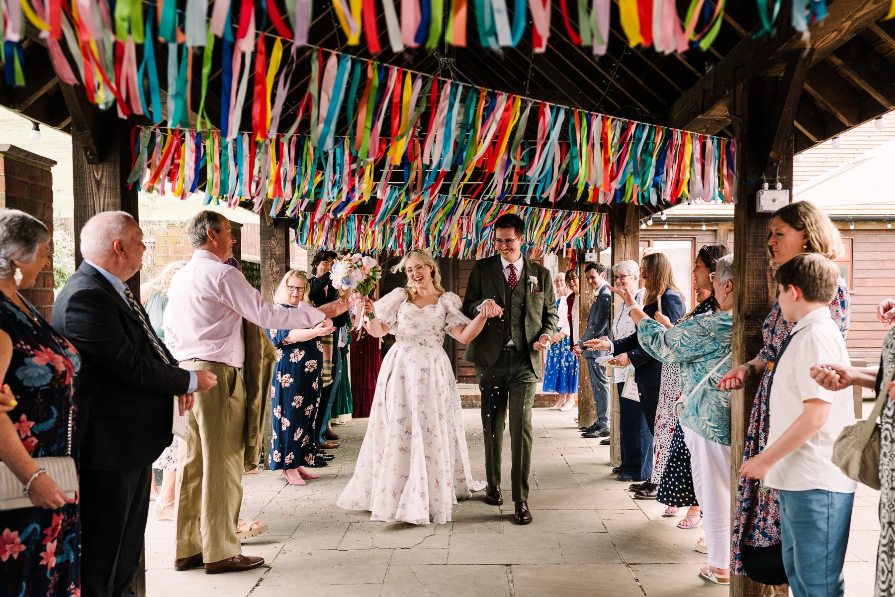 Newly married couple walk through a confetti arch under colourful ribbon garlands at their Long Furlong Barn wedding